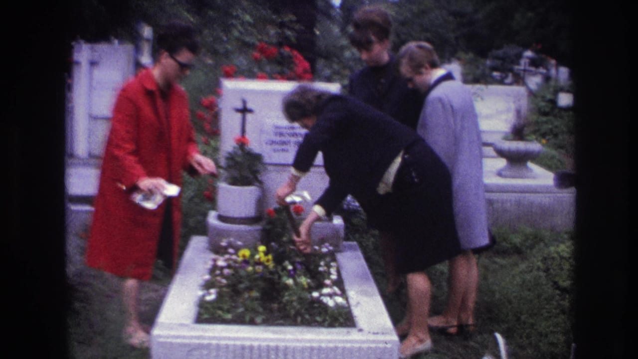 France 1967 8mm frame of four women at a white cemetery grave as one tends flowers beside a cross and tombstone.