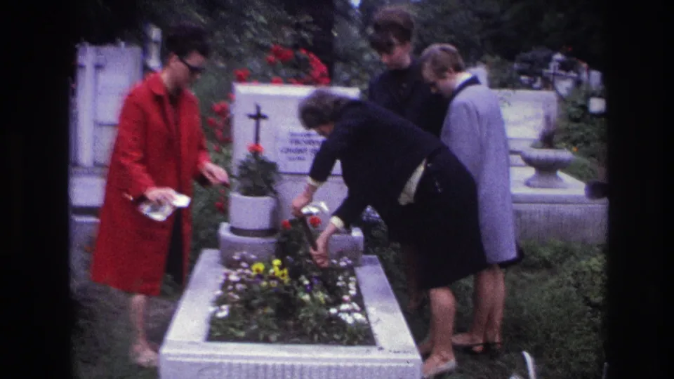 France 1967 8mm frame of four women at a white cemetery grave as one tends flowers beside a cross and tombstone.