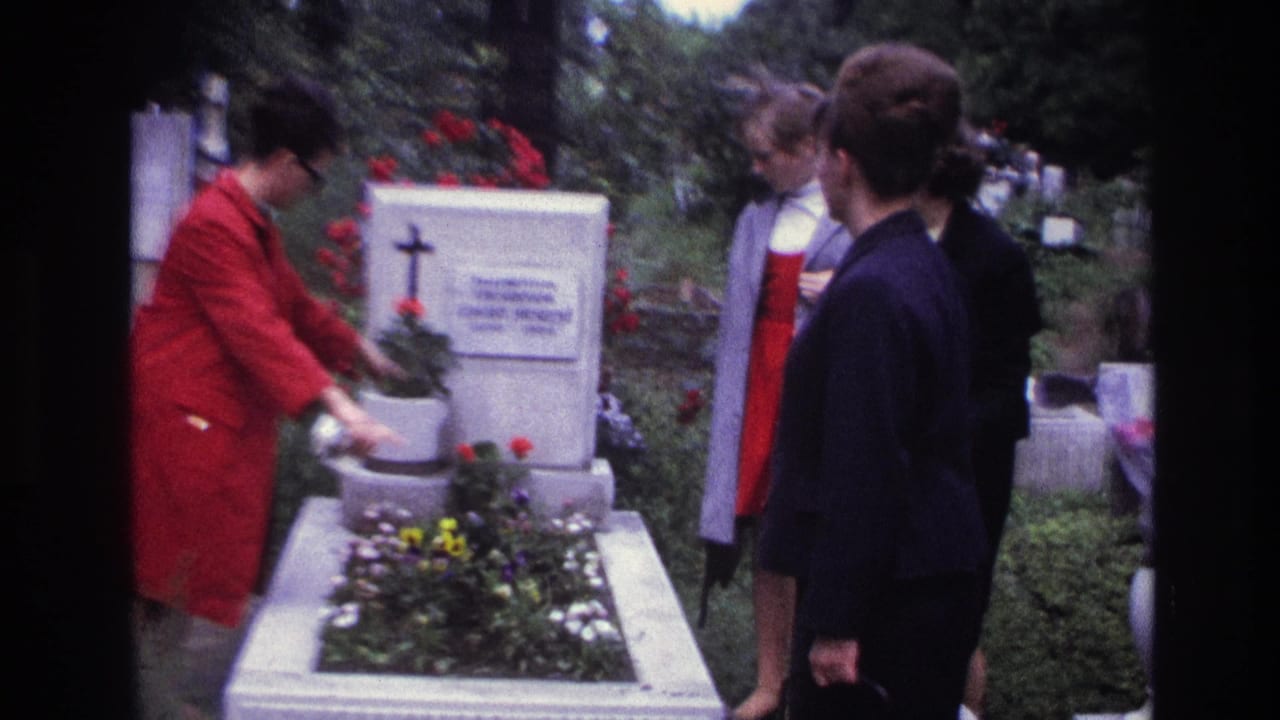 Woman in a red coat gestures near a flowered grave as three women watch.