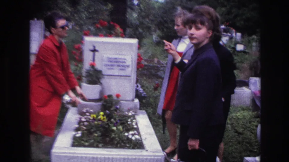 France 1967 8mm frame of women standing at a cemetery grave with flowers, with one woman turning toward the camera.