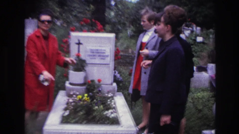 France 1967 8mm frame showing women beside a white flowered grave with a cross and surrounding cemetery greenery.