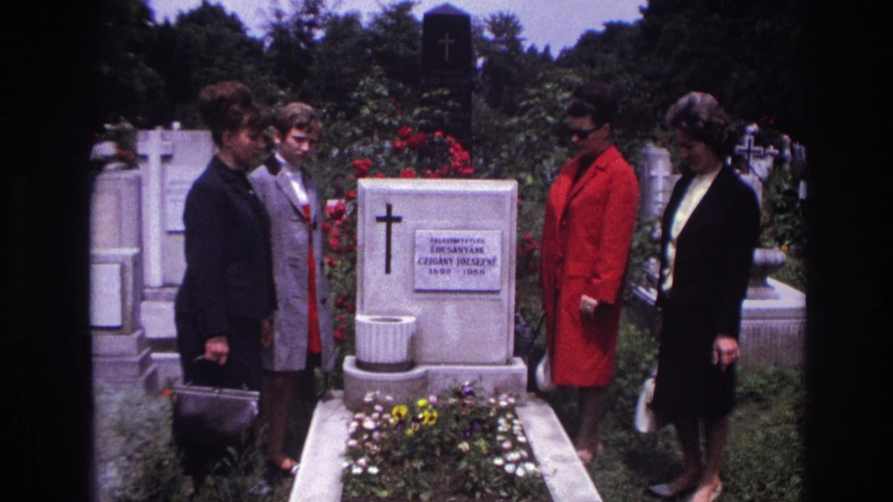 In 1967 France, four adult women stand solemnly beside a white cemetery tombstone with a cross, flowers, and surrounding headstones.