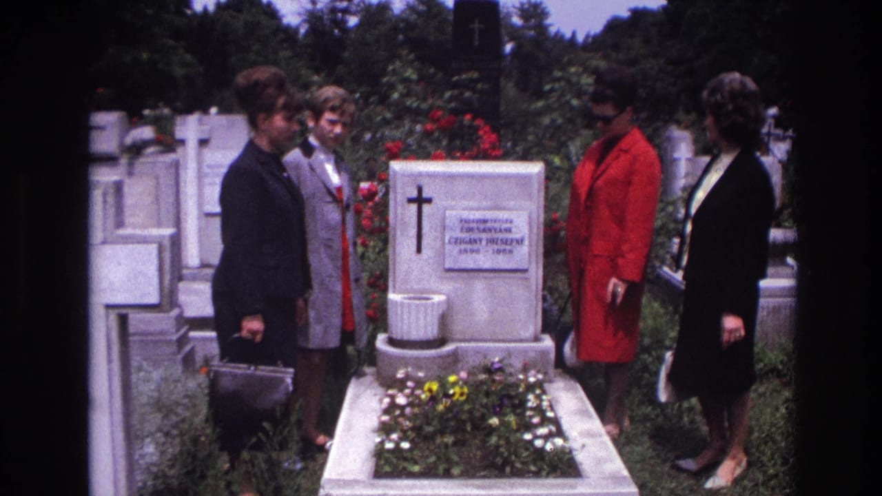 Women in dark clothing and a red coat stand beside a white tombstone with flowers.