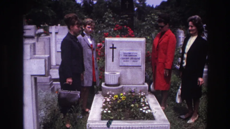 Adult women stand in a French cemetery beside a white grave marker with a cross and Hungarian text, framed by headstones and flowers.