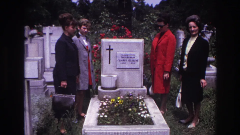 A home-movie frame from 1967 France shows four women gathered beside a flowered grave, with a white tombstone and nearby headstones visible.