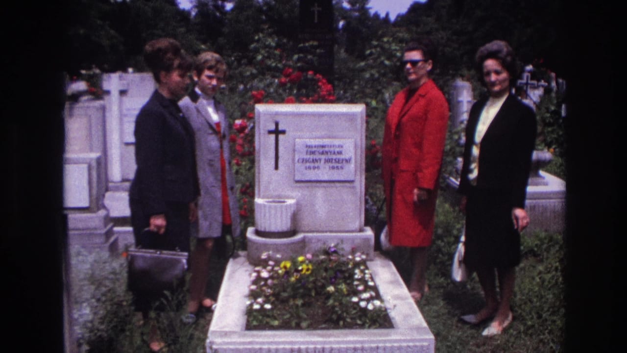 Four women in 1960s dress stand solemnly by a white cemetery grave in France, with flowers and a cross marker visible.