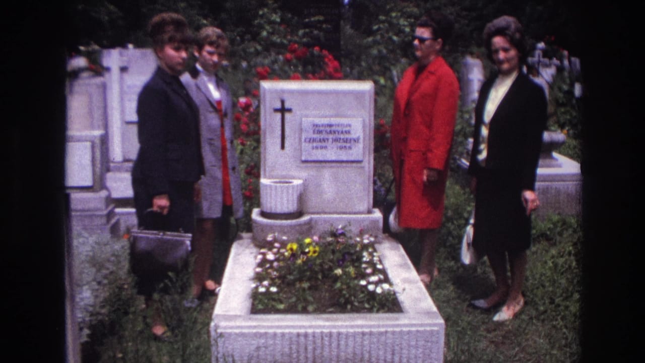Women stand beside a white headstone and flower bed in a cemetery.