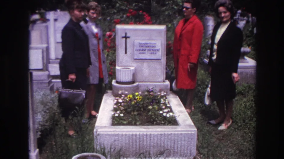 Four women, including one in a red coat, stand solemnly at a flower-adorned grave in a French cemetery setting.