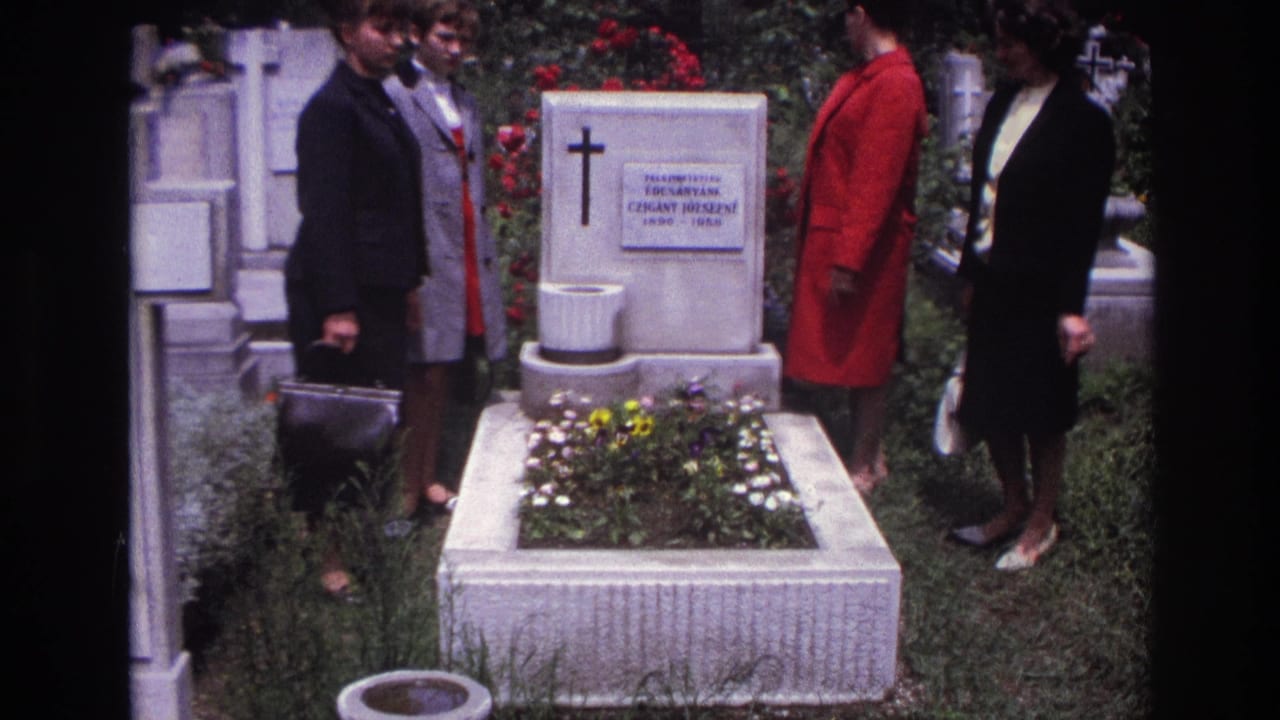 In 1967 France, four women in coats and dresses stand beside a white stone grave with a cross, plaque, flowers, and roses behind it.