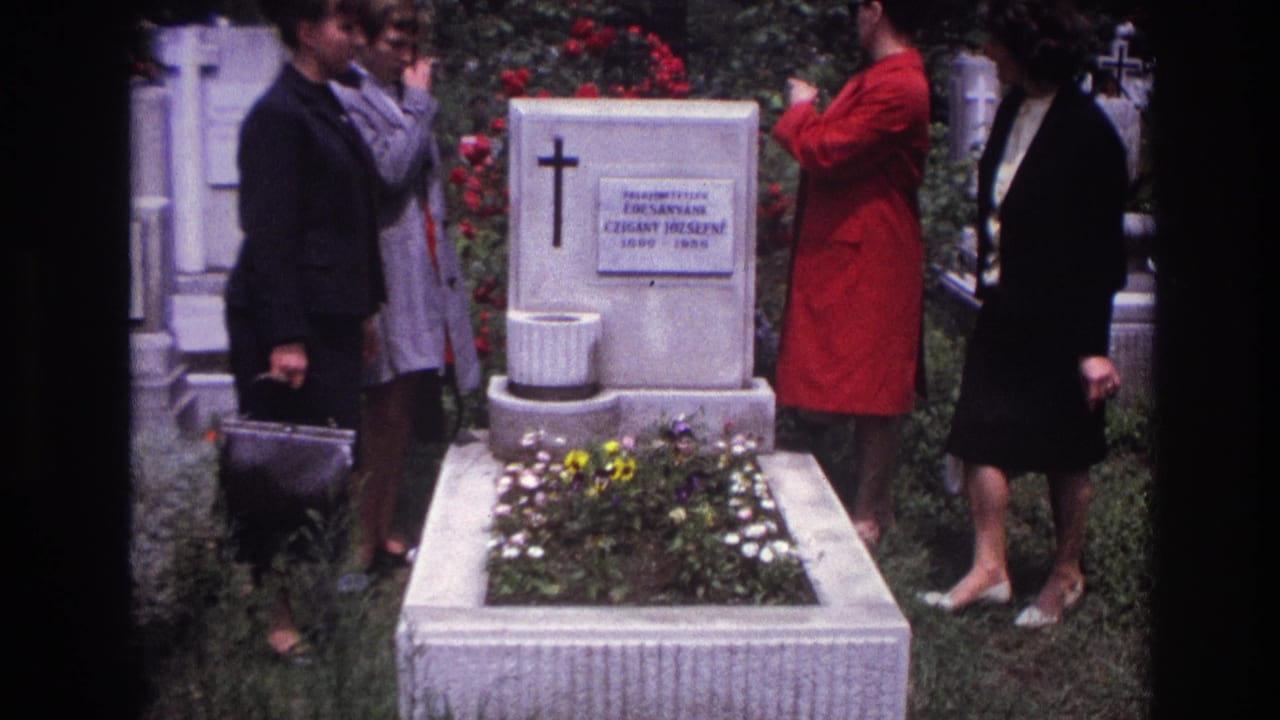 Women in dark and red coats stand beside a white grave with a cross and flower bed.