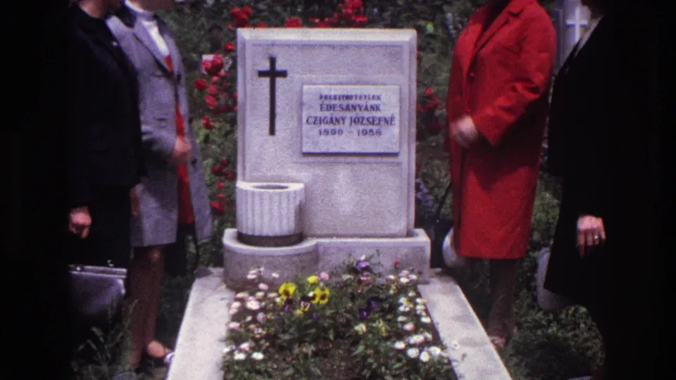 A closer 8mm frame shows the white grave marker with cross and inscription, planted flowers below, and women standing at either side.