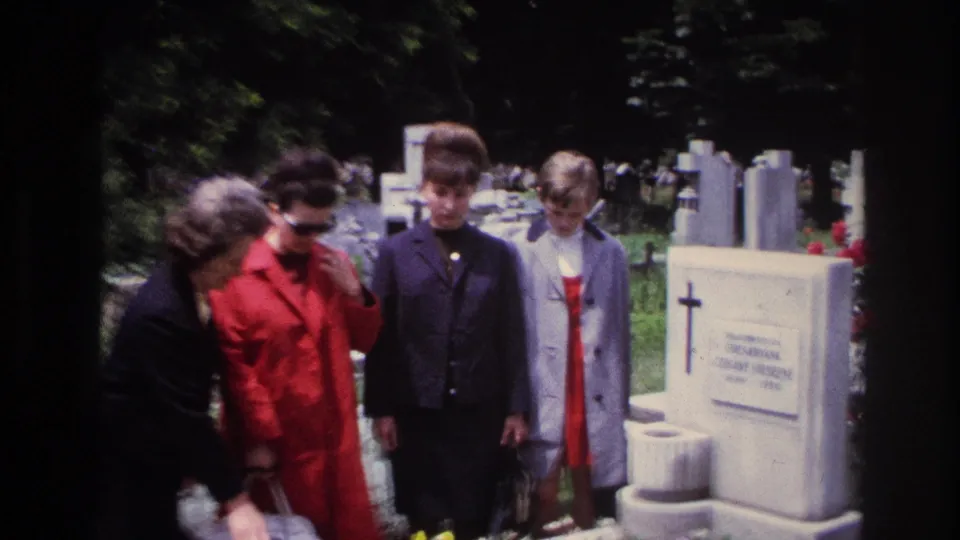 1967 France home-movie frame of four women in period coats standing beside a cemetery grave, with other markers and crosses in the background.