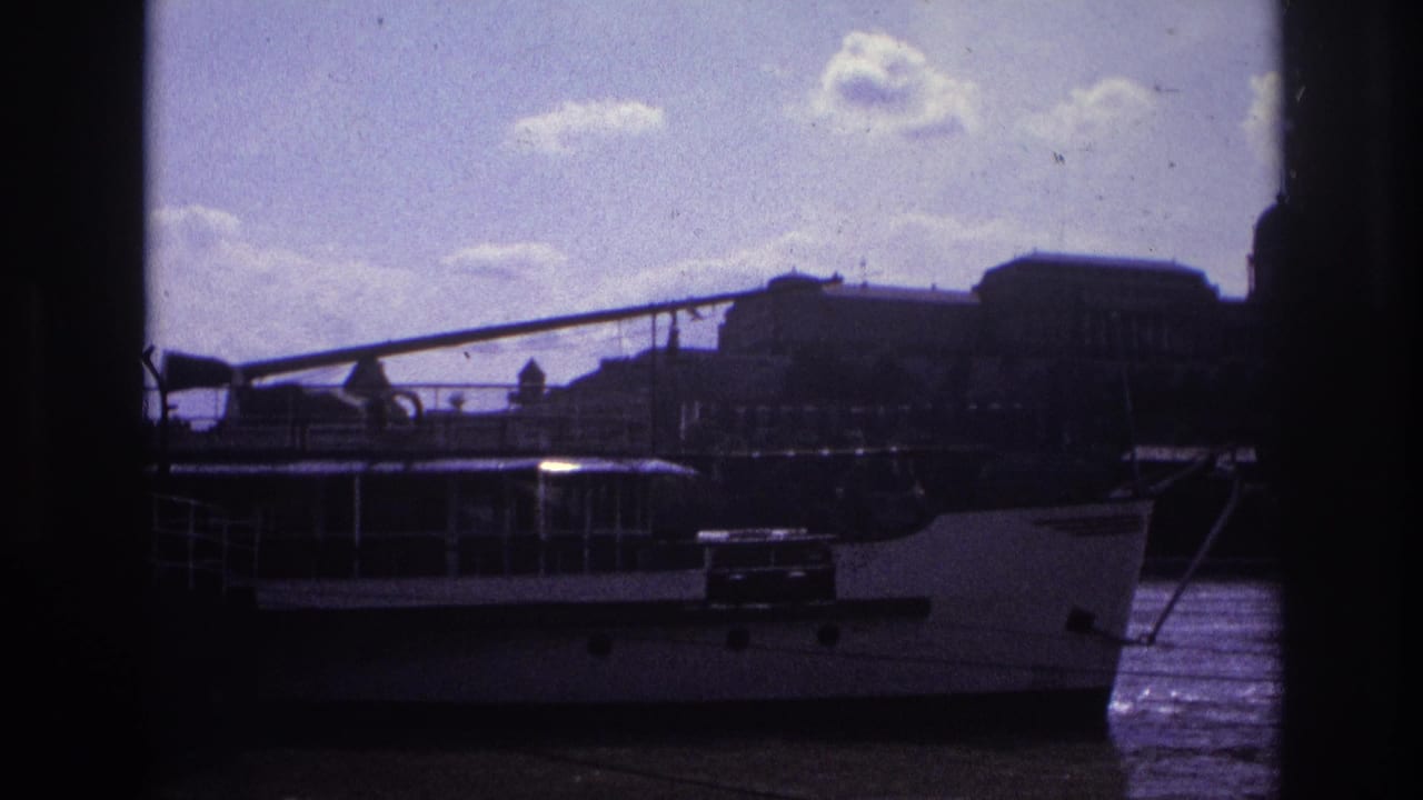 1967 France home-movie frame of a docked riverboat, waterfront structures, and a bright clouded sky with dark scan edges.
