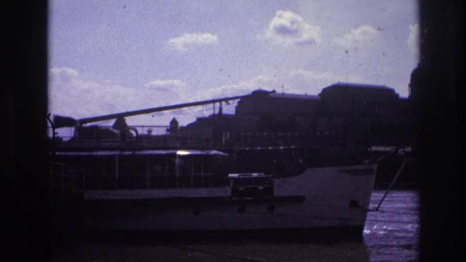 1967 France home-movie frame of a docked riverboat, waterfront structures, and a bright clouded sky with dark scan edges.