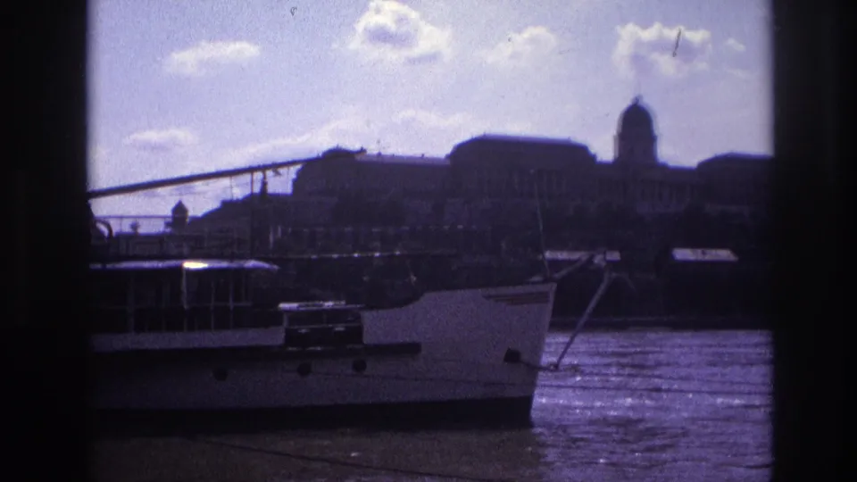 1967 France home-movie frame showing a docked riverboat, shimmering water, and a distant domed skyline under scattered clouds.
