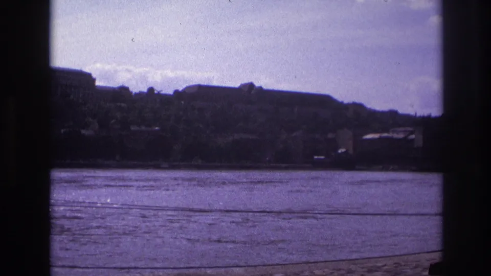 1967 France home-movie view of rippled water and shadowed riverside buildings seen from behind dark vertical supports.