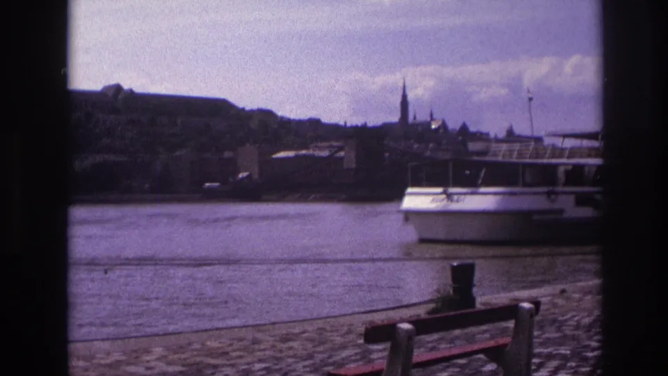 1967 France 8mm frame of a white river tour boat passing a waterfront, with benches, a quay, hillside buildings, and church spires beyond.