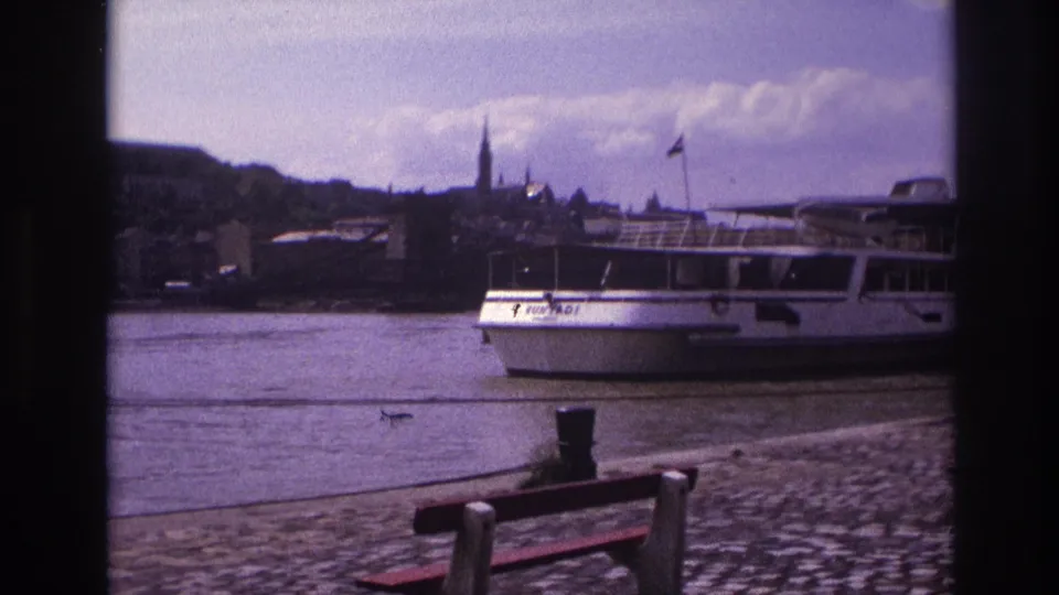1967 France 8mm frame showing a longer view of the white tourist boat beside the cobblestone promenade, with benches and historic buildings behind.