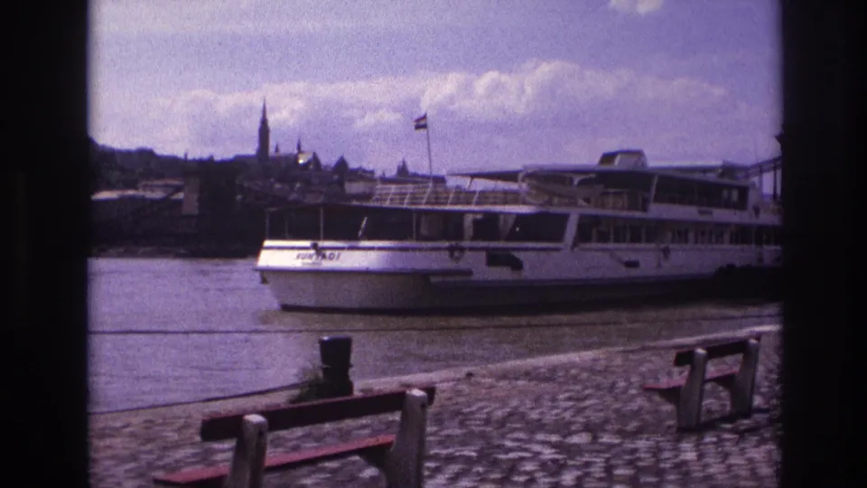 1967 France 8mm frame of a white passenger boat moving along the river past foreground benches, with a historic waterfront skyline in the distance.