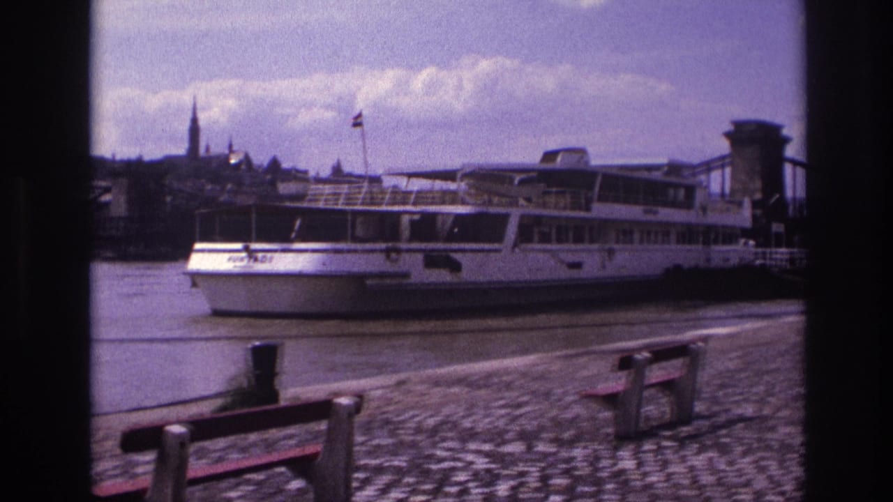 1967 8mm view in France of a docked white passenger boat near a cobblestone riverside and chain suspension bridge.