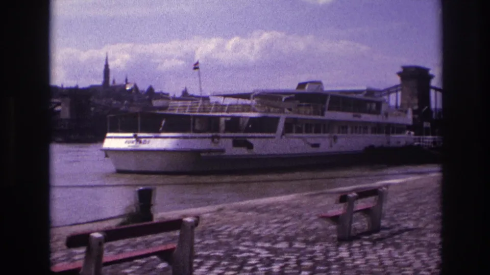 1967 8mm view in France of a docked white passenger boat near a cobblestone riverside and chain suspension bridge.