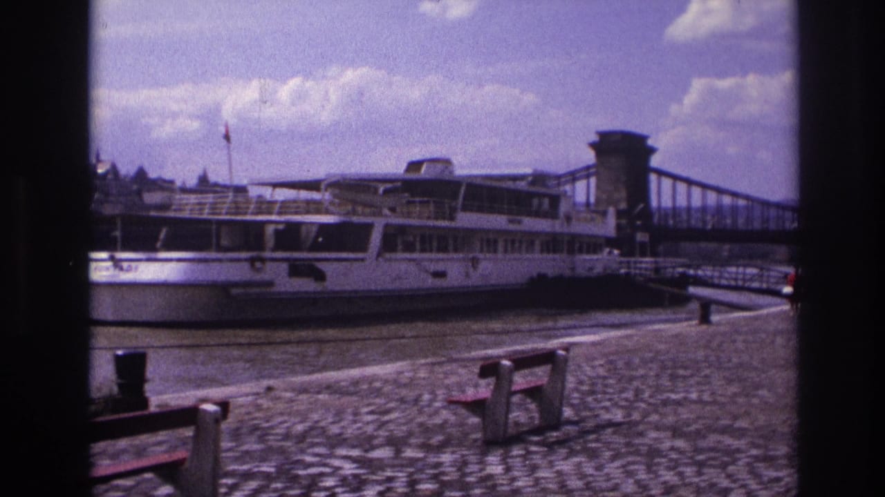 Passenger boat and chain bridge seen from a cobblestone riverside with benches in the foreground.