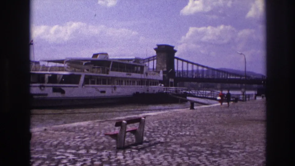 1967 8mm riverside view showing a moored passenger boat, chain bridge, cobblestones, benches, and distant walkers.