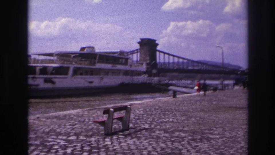 Soft 8mm frame of a French riverside quay with a white passenger boat, chain bridge, bench, and tiny pedestrians in view.
