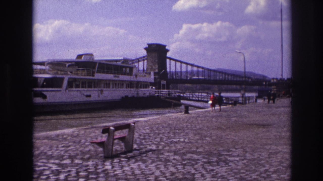 1967 France home-movie frame showing a docked white riverboat, cobblestone promenade, bench, pedestrians, and suspension bridge.