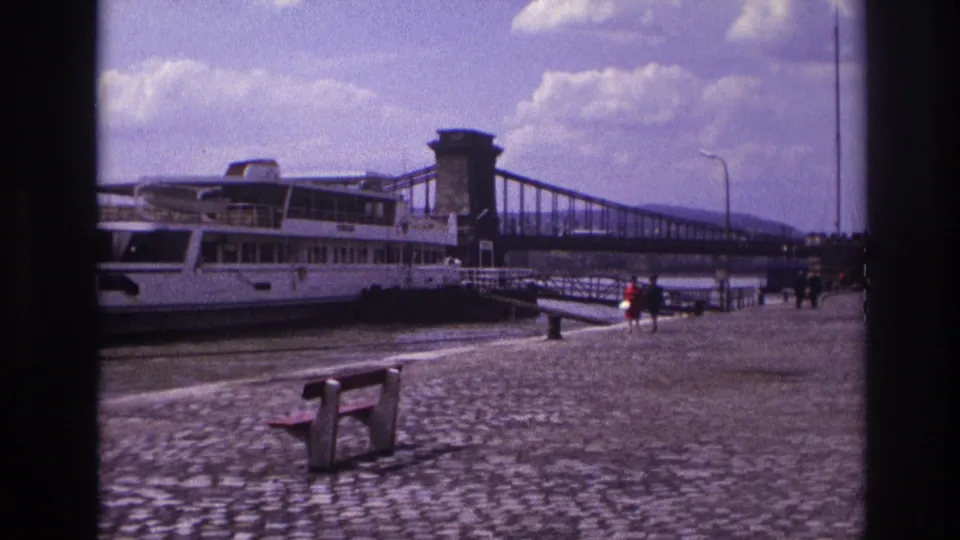 1967 France home-movie frame showing a docked white riverboat, cobblestone promenade, bench, pedestrians, and suspension bridge.