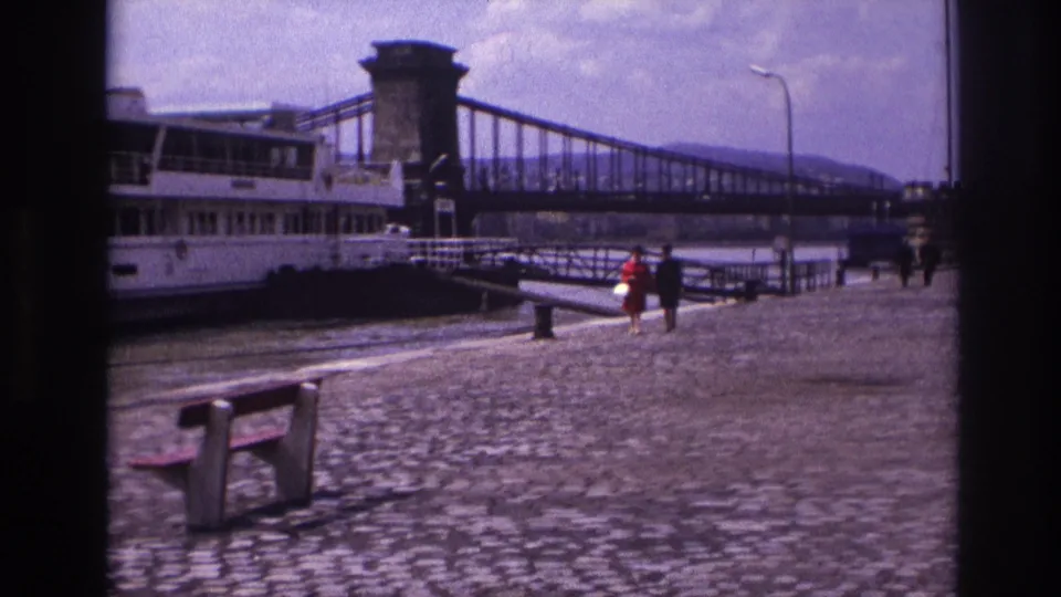 1967 France home-movie view of a cobblestone riverside promenade, bench, moored boat, bridge tower, and distant pedestrians.