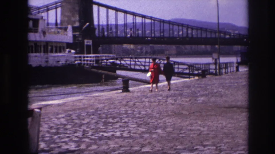 1967 France frame showing a woman in red and a man in dark clothing walking near a riverboat gangway under a suspension bridge.
