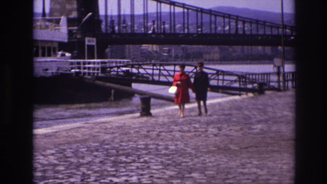 In 1967 France, two women walk along a cobblestone quay near a riverboat and large metal bridge.
