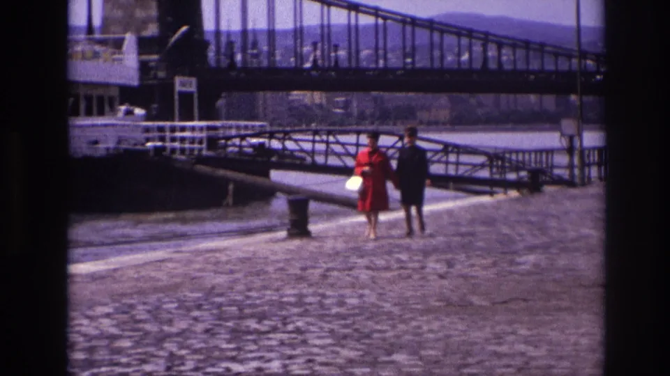 In 1967 France, two women walk along a cobblestone quay near a riverboat and large metal bridge.