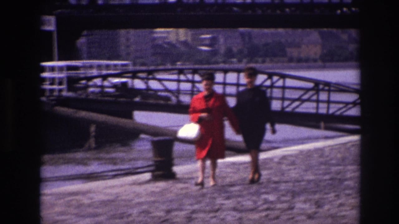 Two women walk closer to the camera beside railings and a metal bridge.