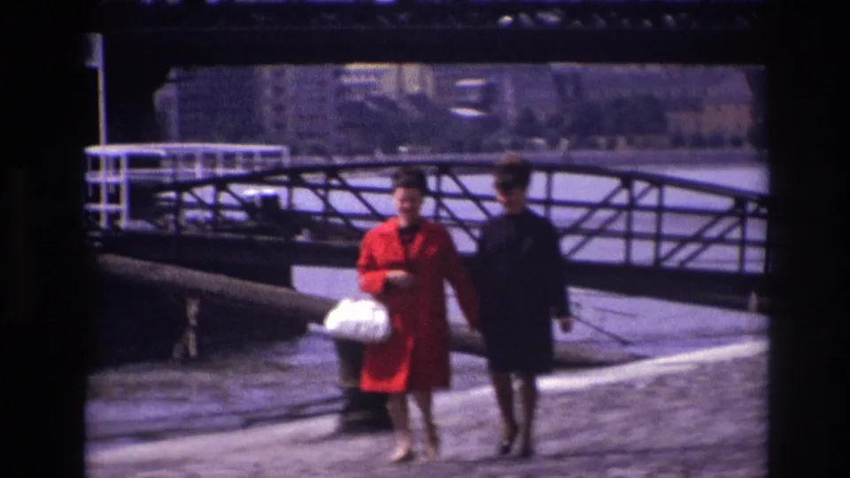 A woman in a red coat and a companion in dark clothing pass the camera along a French riverside quay in 1967.