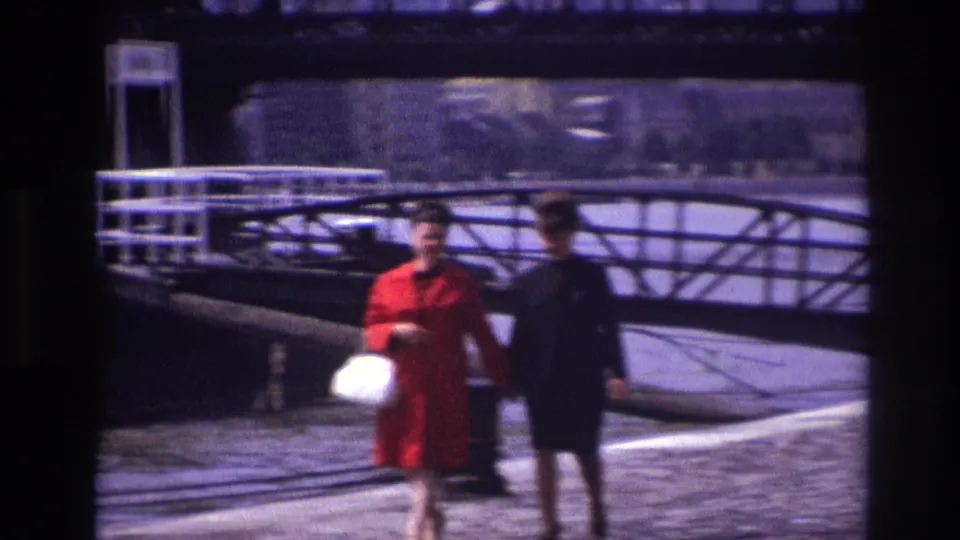 The walkers move out of frame on a 1967 French riverside promenade, backed by bridge rails, water, and distant buildings.