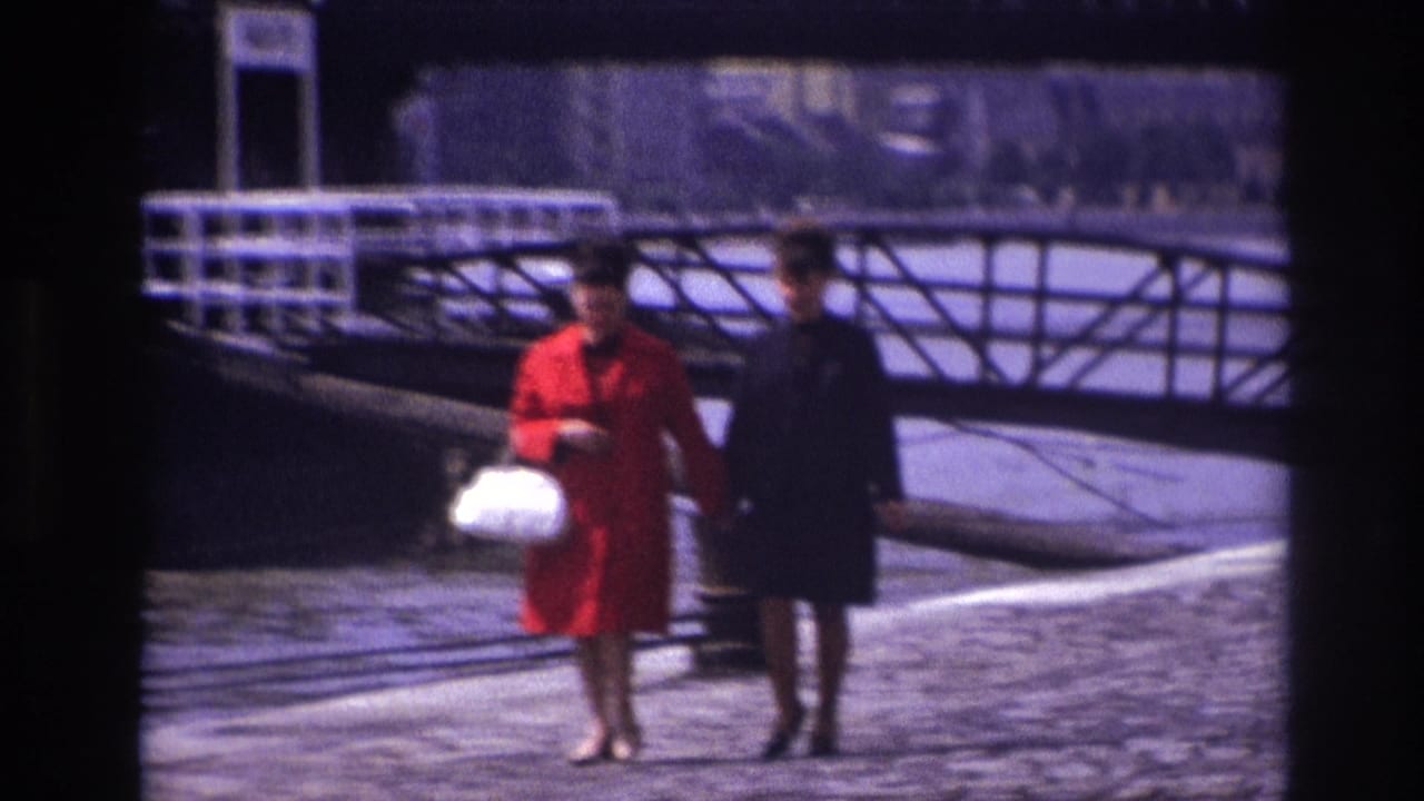 Two women, one in a red coat with a white handbag, walk along a cobblestone riverside promenade in 1967 travel footage.