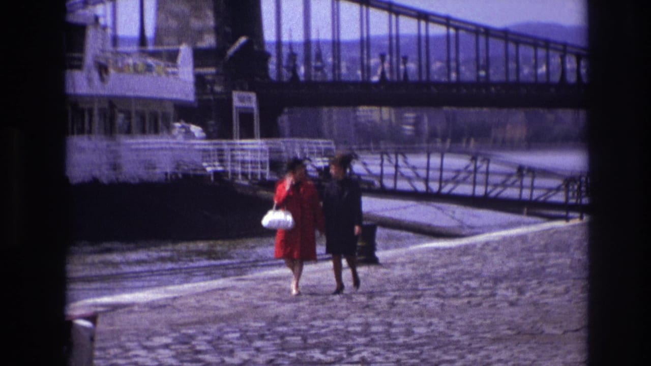 Two women walk farther along the riverside with a large boat and bridge behind them.