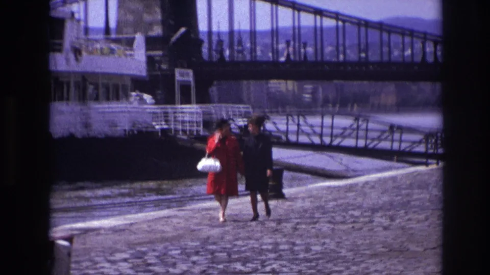A wide 1967 riverside view shows two women walking on cobblestones near a moored riverboat, gangway, and bridge.