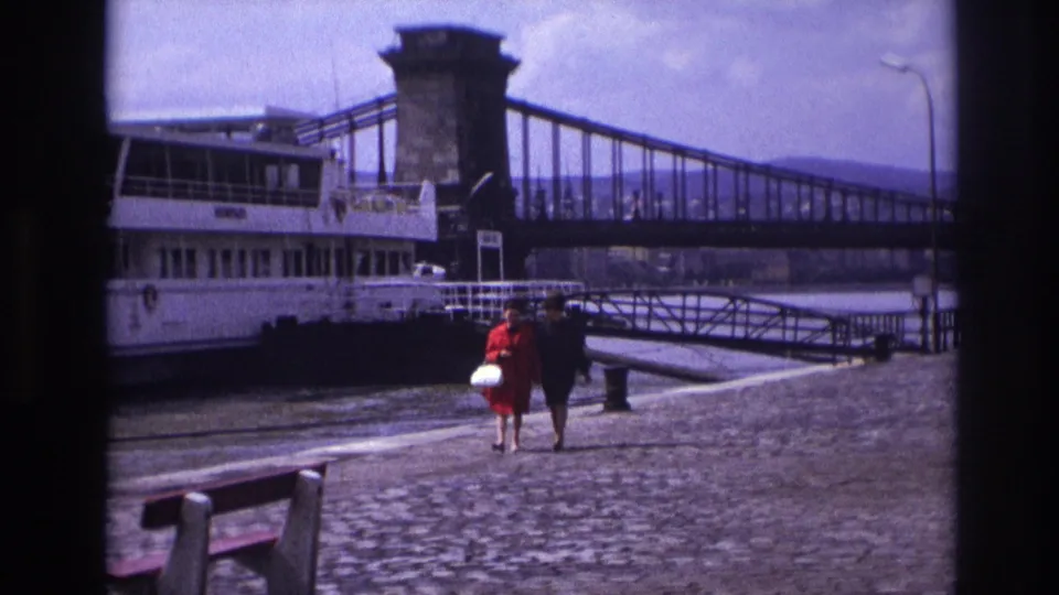 The women appear smaller in a 1967 home-movie frame showing a riverside quay, benches, a boat, and a bridge span.
