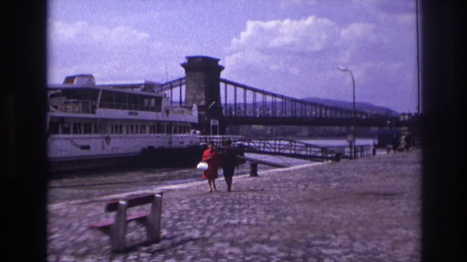 A wide 1967 travel-film frame shows the pair walking away along the riverfront, with benches, a boat, bridge, and open sky.
