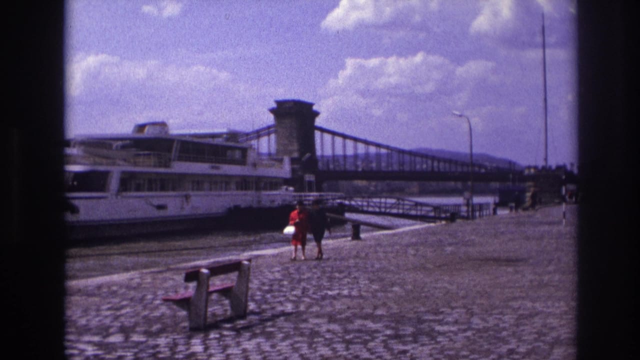 1967 France home-movie frame with two women walking on a cobblestone riverside promenade near a white riverboat and bridge.