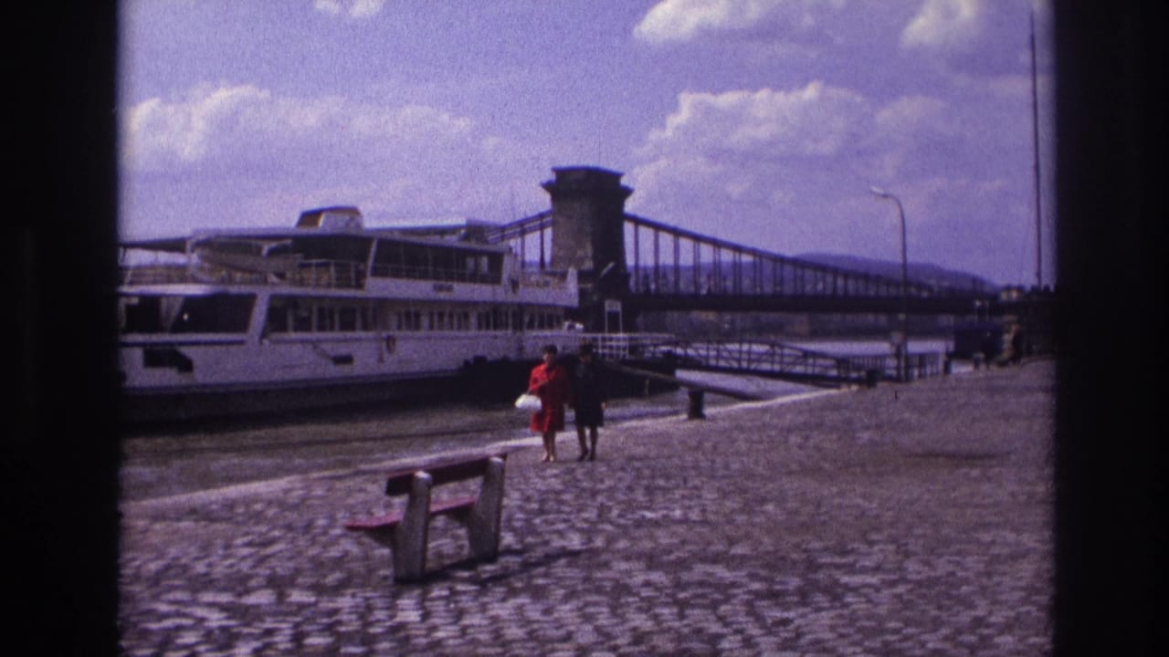 Two women, one in a red dress, walk along a French riverside quay in 1967 with a moored riverboat and suspension bridge visible.