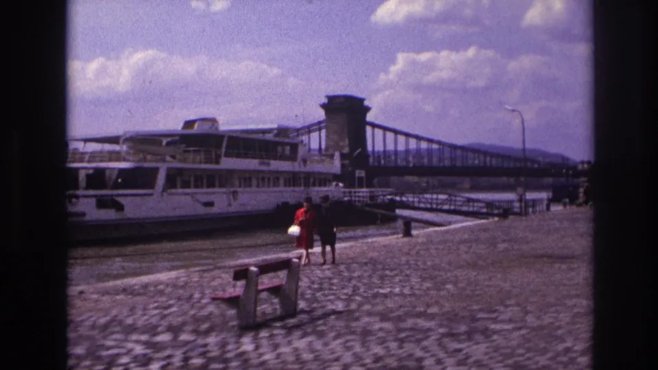 1967 France riverside home-movie view with a white riverboat beside the quay and two women walking past a wooden bench.