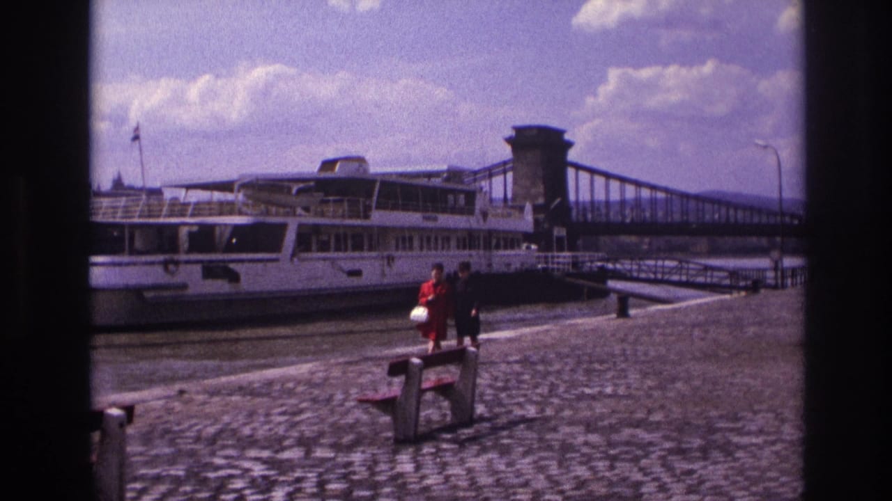In 1967 France, two women stroll along a cobblestone riverside promenade with a large white riverboat and suspension bridge behind them.
