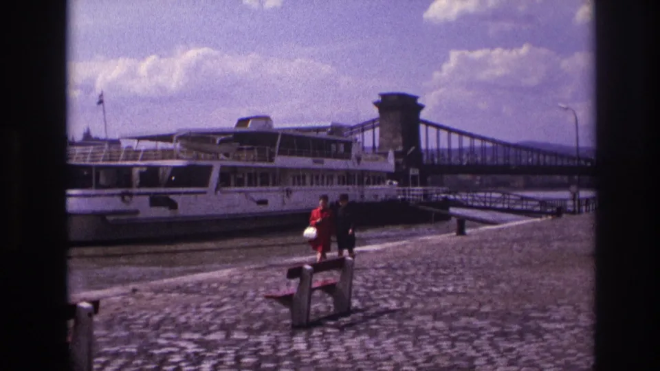 In 1967 France, two women stroll along a cobblestone riverside promenade with a large white riverboat and suspension bridge behind them.