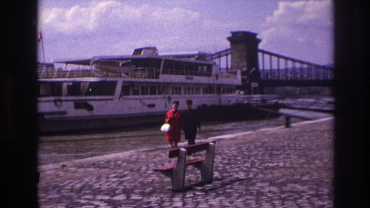 Two women continue walking near a bench with the riverboat and bridge in the background.