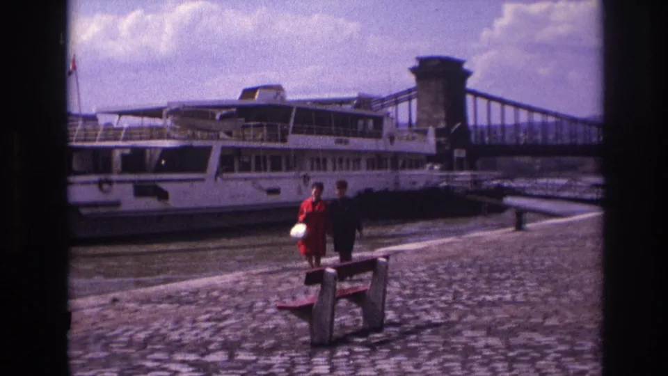 The two women move closer along the French riverside quay, passing a bench as the docked white riverboat fills the background.
