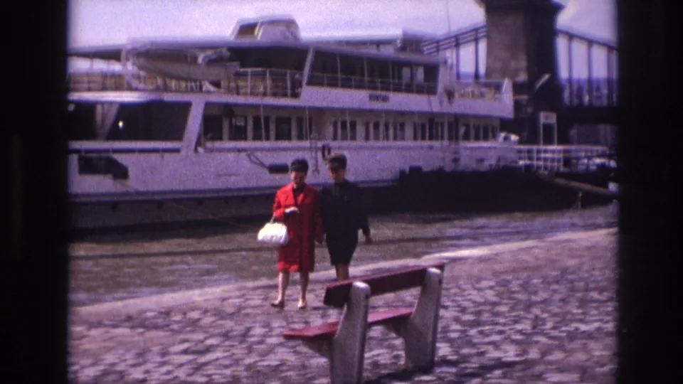 Two women, one in a red coat carrying a white handbag, walk side by side on a cobblestone quay before a white riverboat in France.